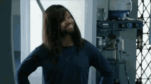 a man with long hair and a beard is standing with his hands on his hips in a room .