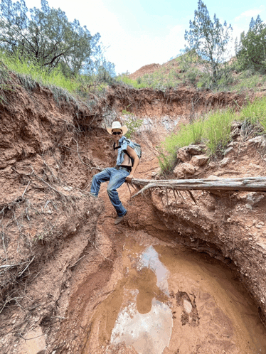 a man in a cowboy hat stands on a log over a muddy river