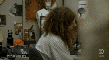 a man with long curly hair sits at a desk with a comedy central logo
