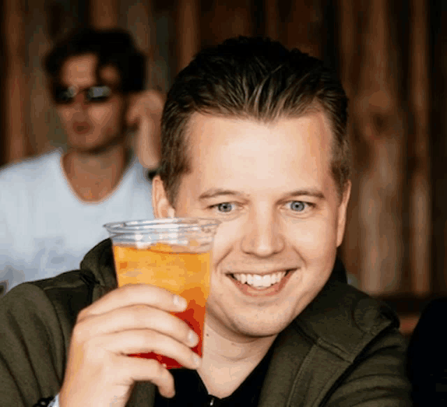 a man in a green jacket is smiling while holding a glass of orange liquid