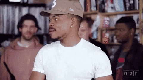 a man wearing a hat and a white t-shirt is standing in front of a group of men .