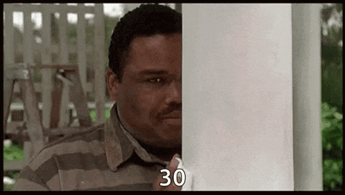 a man in a striped shirt is peeking out from behind a white pole and says `` 30 '' .