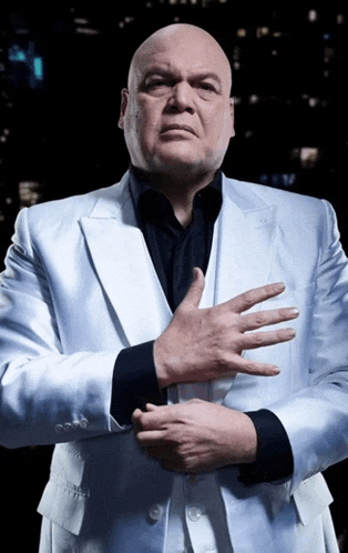 a bald man in a white suit and black shirt holds his hands together