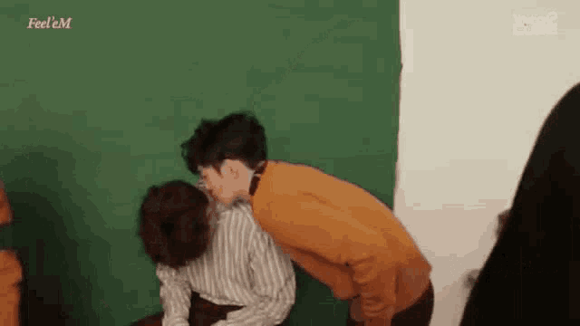 a man in an orange sweater is kissing another man on the cheek in front of a green wall .
