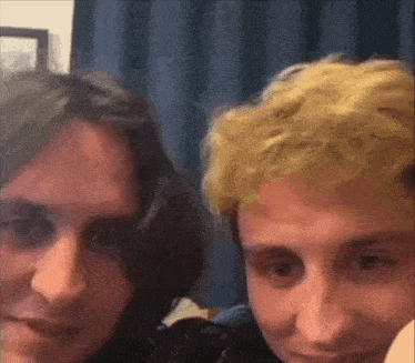 two men are posing for a picture together in a room . one has blonde hair and the other has black hair .
