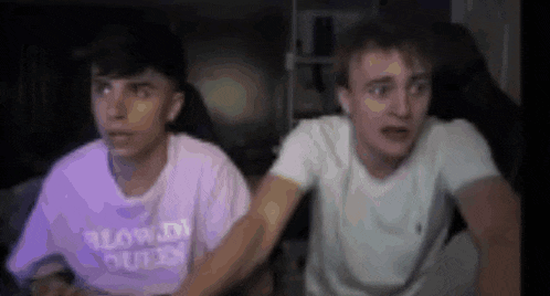 two young men are sitting next to each other in front of a computer screen . one of the men is wearing a purple shirt .
