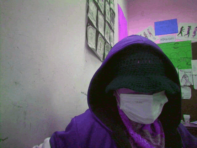 a person wearing a mask and a purple hoodie stands in front of a bulletin board with drawings on it