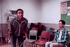 two men are standing in a classroom with chairs and a chalkboard .