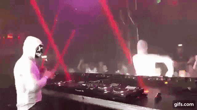 a man in a white hoodie is standing in front of a dj booth in a club .