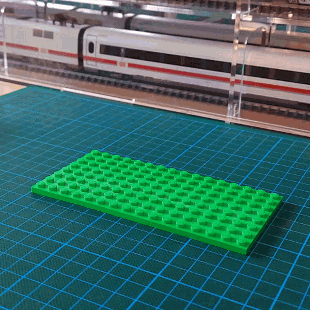 a green lego block sits on a green cutting mat in front of a model train