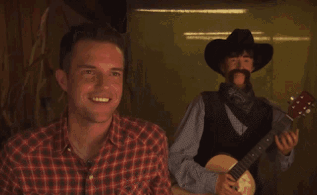 a man in a plaid shirt is smiling while a man in a cowboy hat holds a guitar