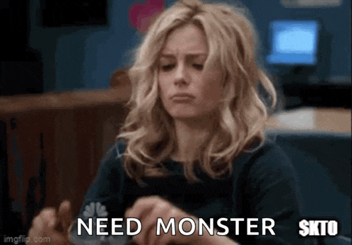 a woman is sitting at a table and making a face while holding her hands together and says `` need monster '' .