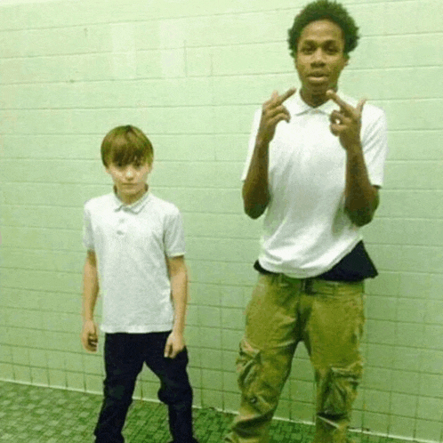 two boys are standing next to each other in front of a green tiled wall and one of them is giving the middle finger
