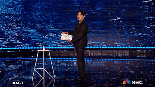 a man in a suit stands on a stage holding a box that says nbc