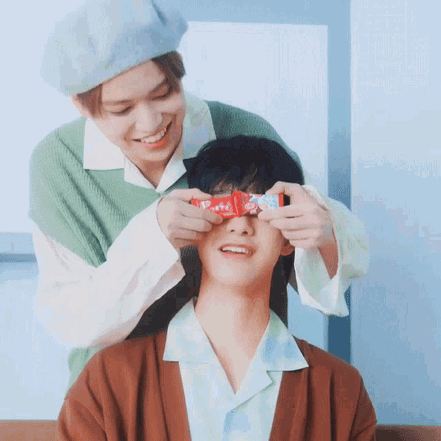 a man is covering another man 's eyes with a box that says ' strawberry ' on it