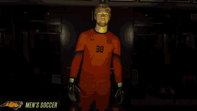a man in an orange under armour shirt stands in a dark locker room with the words men 's soccer below him