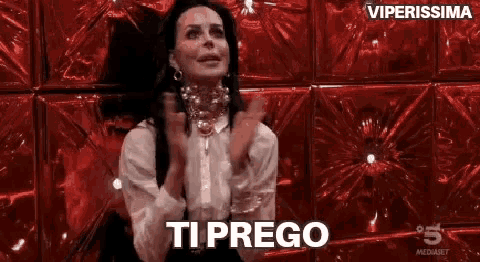 a woman is sitting in front of a red wall with her hands folded and the word ti prego written on it .