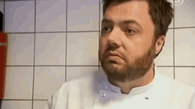 a man with a beard is wearing a white chef 's coat and making a funny face .