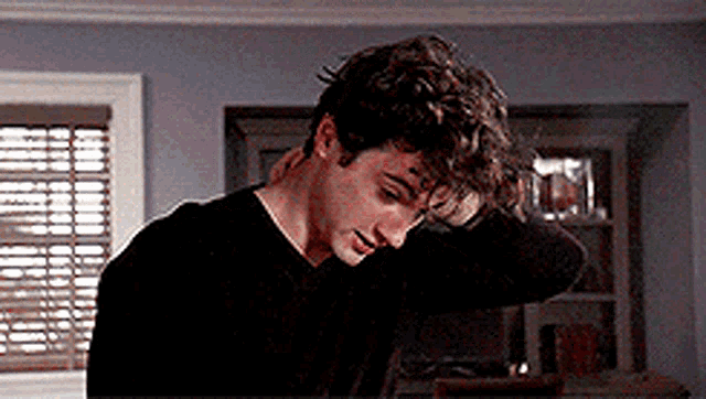 a young man with curly hair is standing in a living room with his hand on his head .