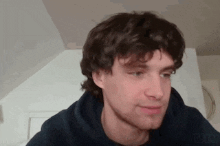 a young man with curly hair and a beard is wearing a hoodie and looking at the camera .