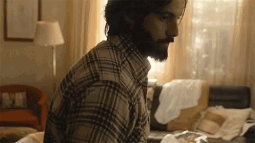 a man with a beard is sitting on a bed in a living room wearing a plaid shirt and glasses .