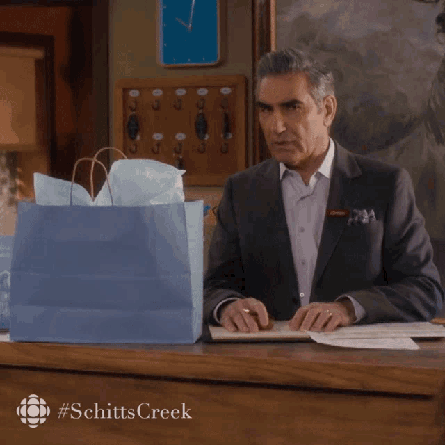 a man in a suit sits at a desk in front of a sign that says schitts creek on it