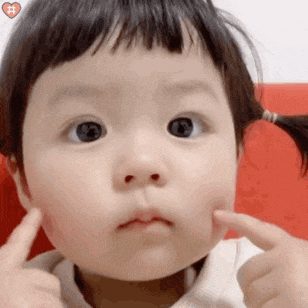 a baby girl with pigtails is making a funny face and pointing at her cheek .