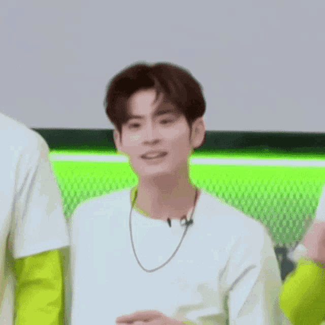 a young man wearing a white t-shirt and a necklace is smiling while standing in front of a green wall .