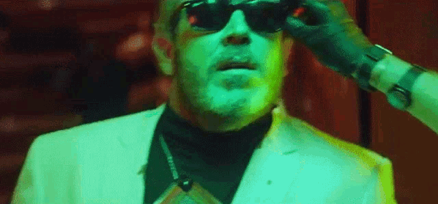 a man wearing sunglasses and a white suit is being adjusted by a person in a green light .