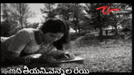a black and white photo of a woman laying on the grass writing on a piece of paper .