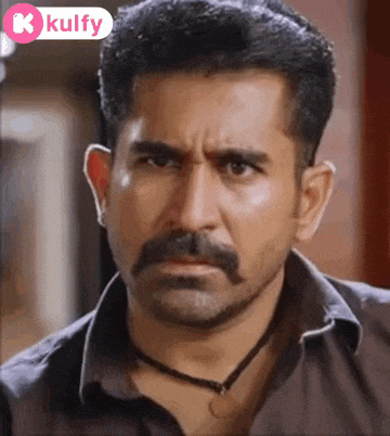 a man with a mustache and a necklace is looking at the camera .