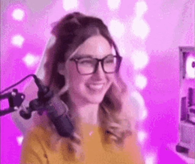 a woman wearing glasses is smiling in front of a microphone in a pink room .