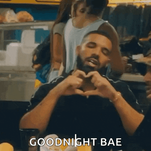a man with a beard is sitting at a table with his hands in the shape of a heart and says goodnight bae .