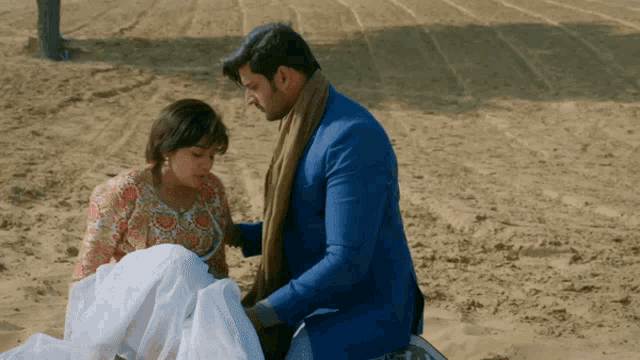 a man in a blue suit is helping a woman in a white dress in the sand
