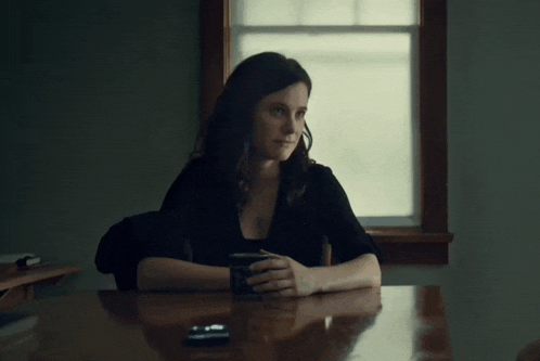 a woman sits at a table in front of a window holding a cup of coffee