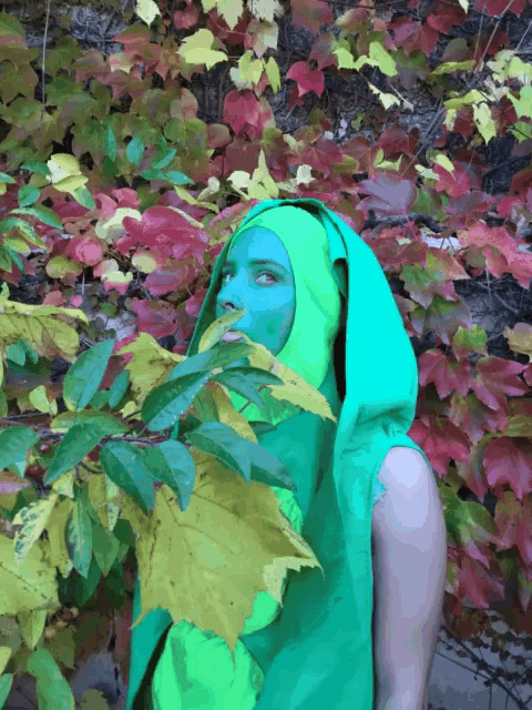 a person with green paint on their face is standing in front of leaves