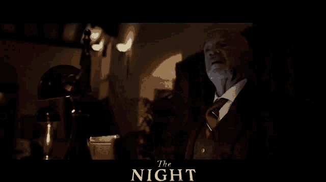 a man in a suit and tie sits in a dark room with the words the night written above him
