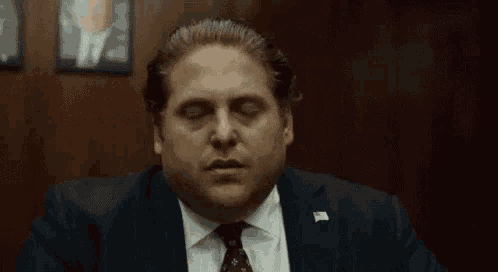 a man in a suit and tie is sitting in a dark room with his eyes closed and making a funny face .