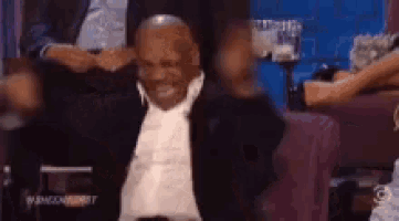 a man in a suit is sitting in a chair with his arms outstretched and a woman behind him .