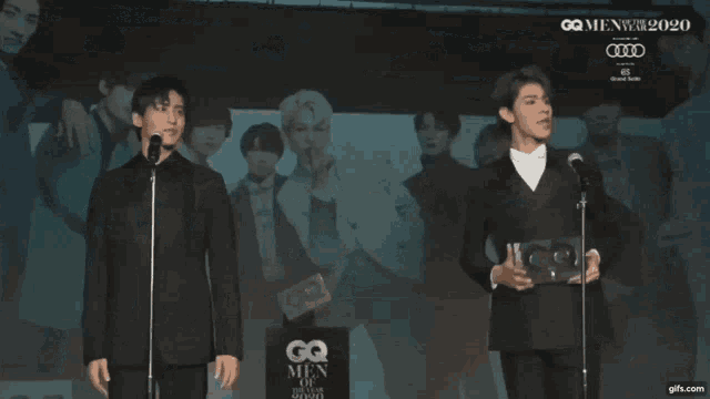 a man in a suit stands in front of a large screen that says gq men of the year 2020