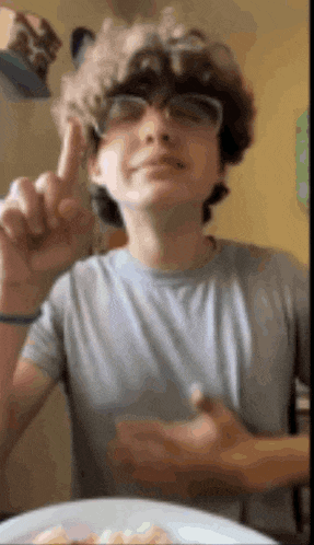 a young man wearing glasses is giving the middle finger while sitting at a table with a plate of food .