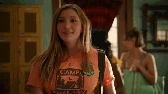 a girl wearing an orange t-shirt with a camp logo on it is standing in a room .