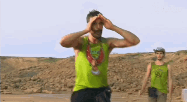 a man in a green tank top is covering his eyes with his hands while another man stands behind him .