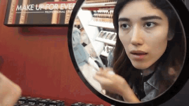 a woman looking at her face in a mirror with a make up for ever sign behind her