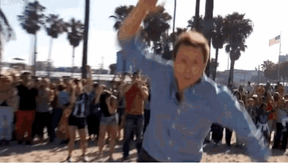a man in a blue shirt is dancing in front of a crowd on the beach