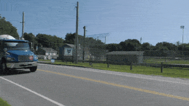 a blue truck is driving down a road in front of a baseball field with a sign that says ' a.m. '