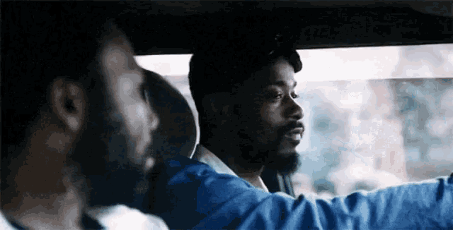 a man with a beard is driving a car with another man sitting in the back seat .