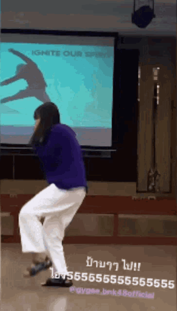 a woman in a purple shirt and white pants is dancing in front of a screen that says ignite our spirit