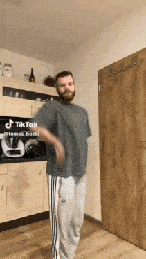 a man in a grey shirt and white pants is dancing in a kitchen with a tiktok hashtag