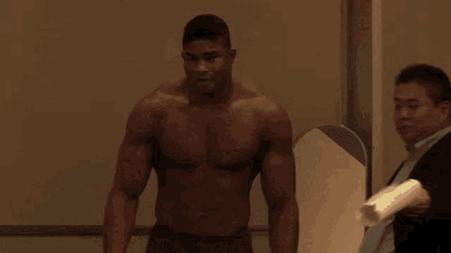 a man without a shirt is walking in a hallway with a green exit sign above him